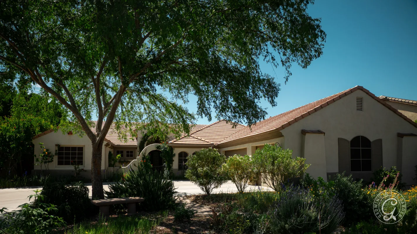 Single-story house with a tile roof, large tree, shrubs, and garden in the front yard on a sunny day.