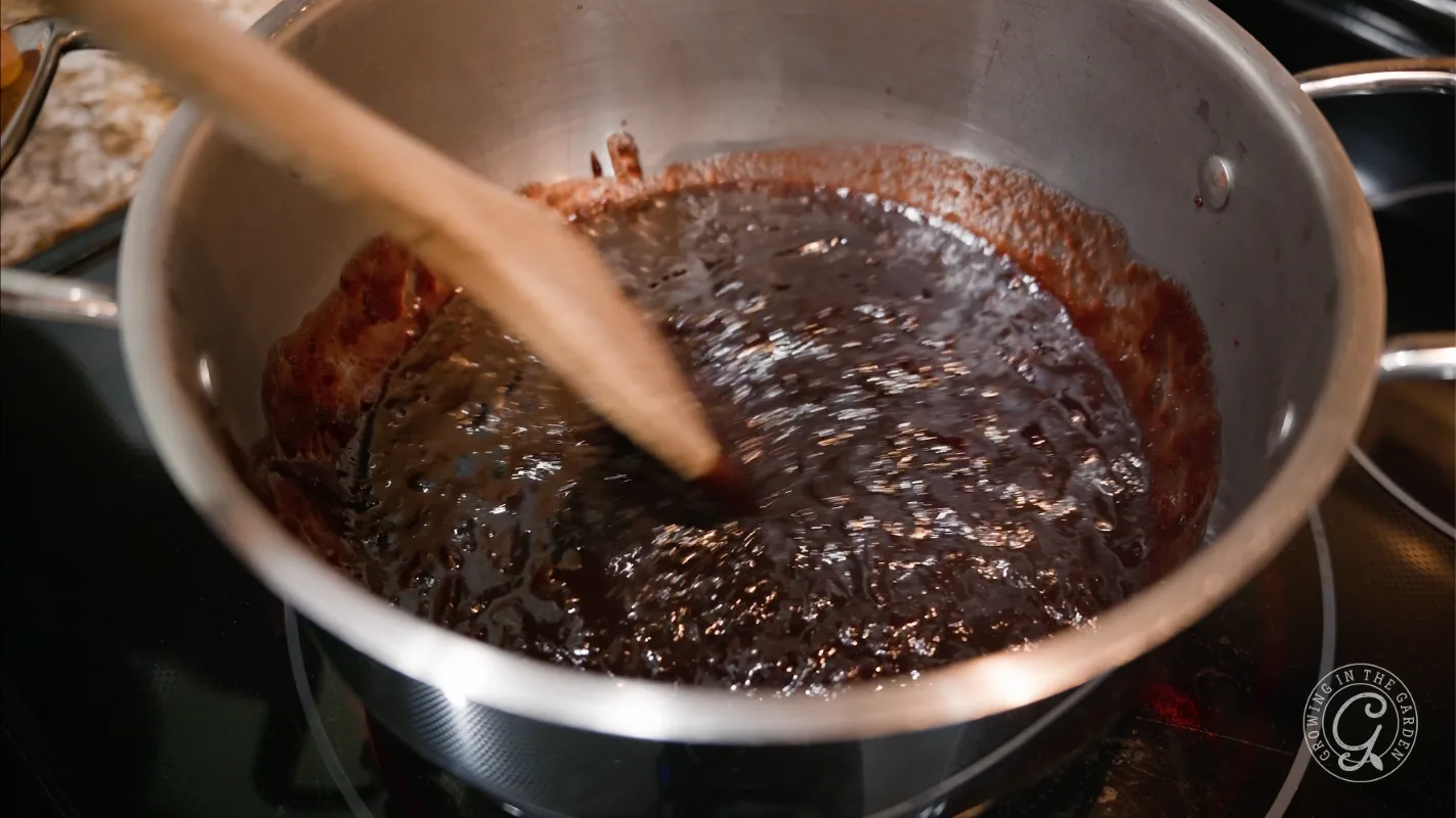 A wooden spoon stirs a dark, thick sauce in a stainless steel pot on a stove.