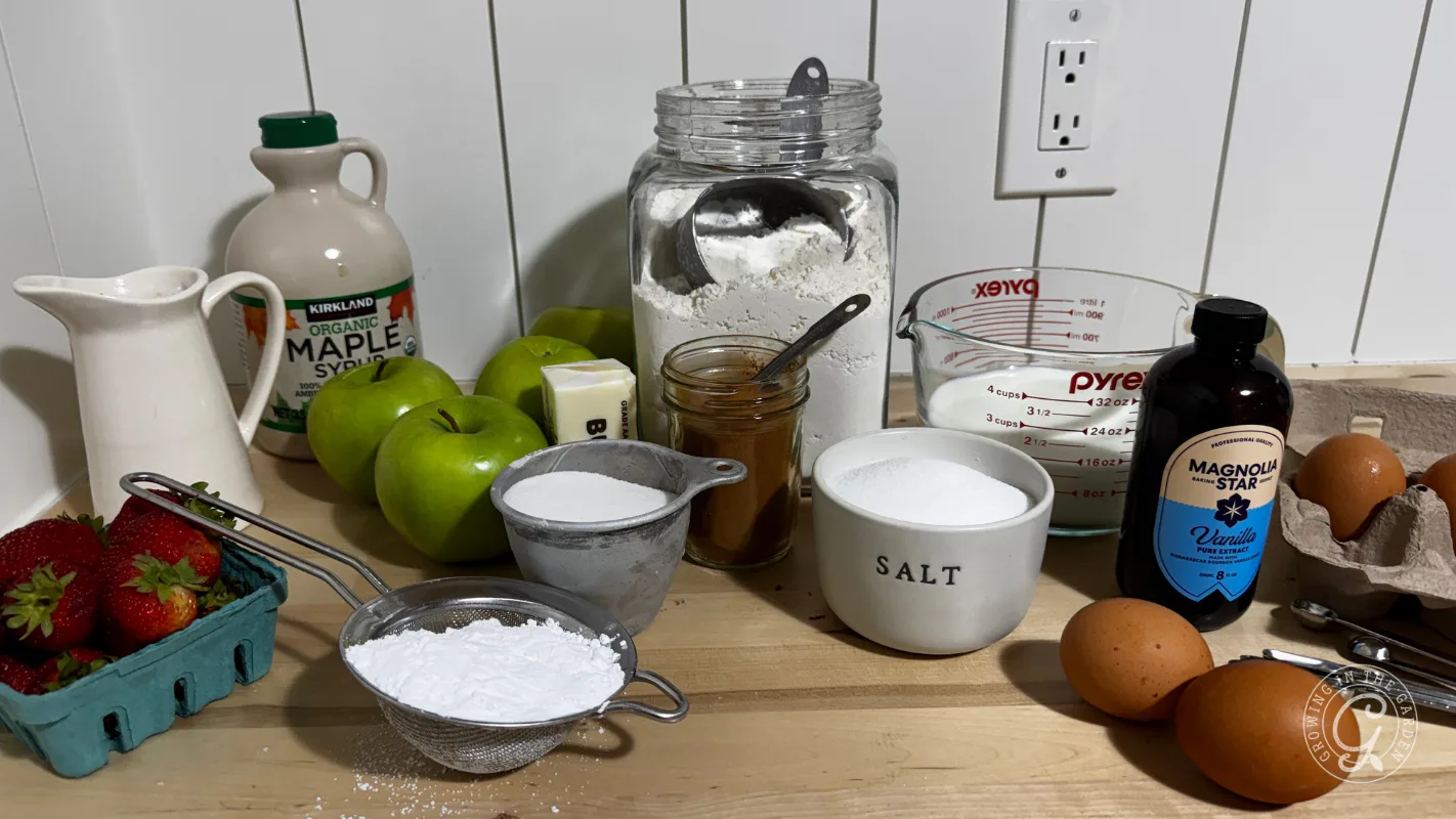 Baking ingredients on a counter: apples, strawberries, eggs, flour, syrup, sugar, salt, butter, and milk.
