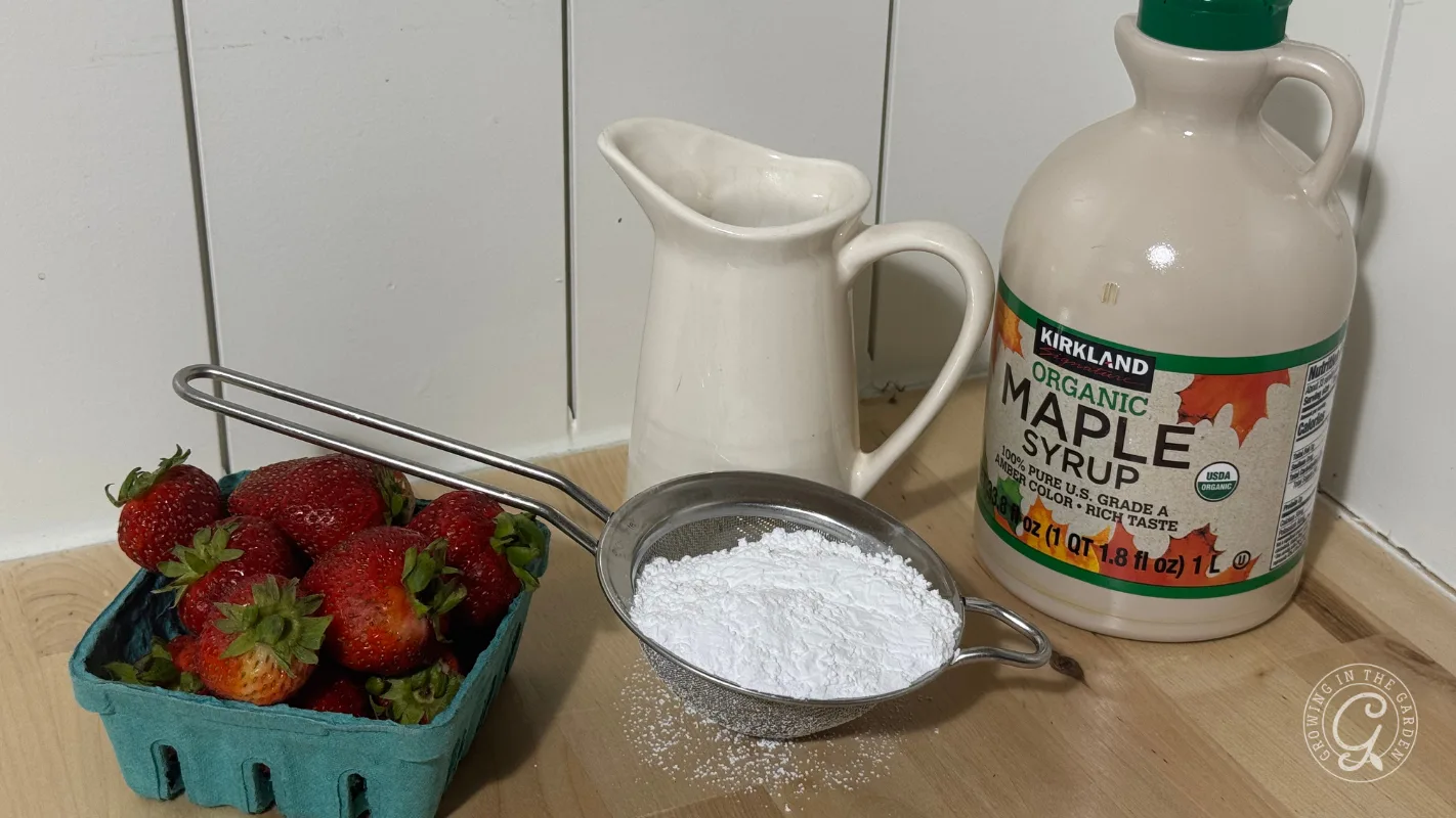 Basket of strawberries, powdered sugar in a strainer, syrup bottle, and a ceramic pitcher on a wooden counter.
